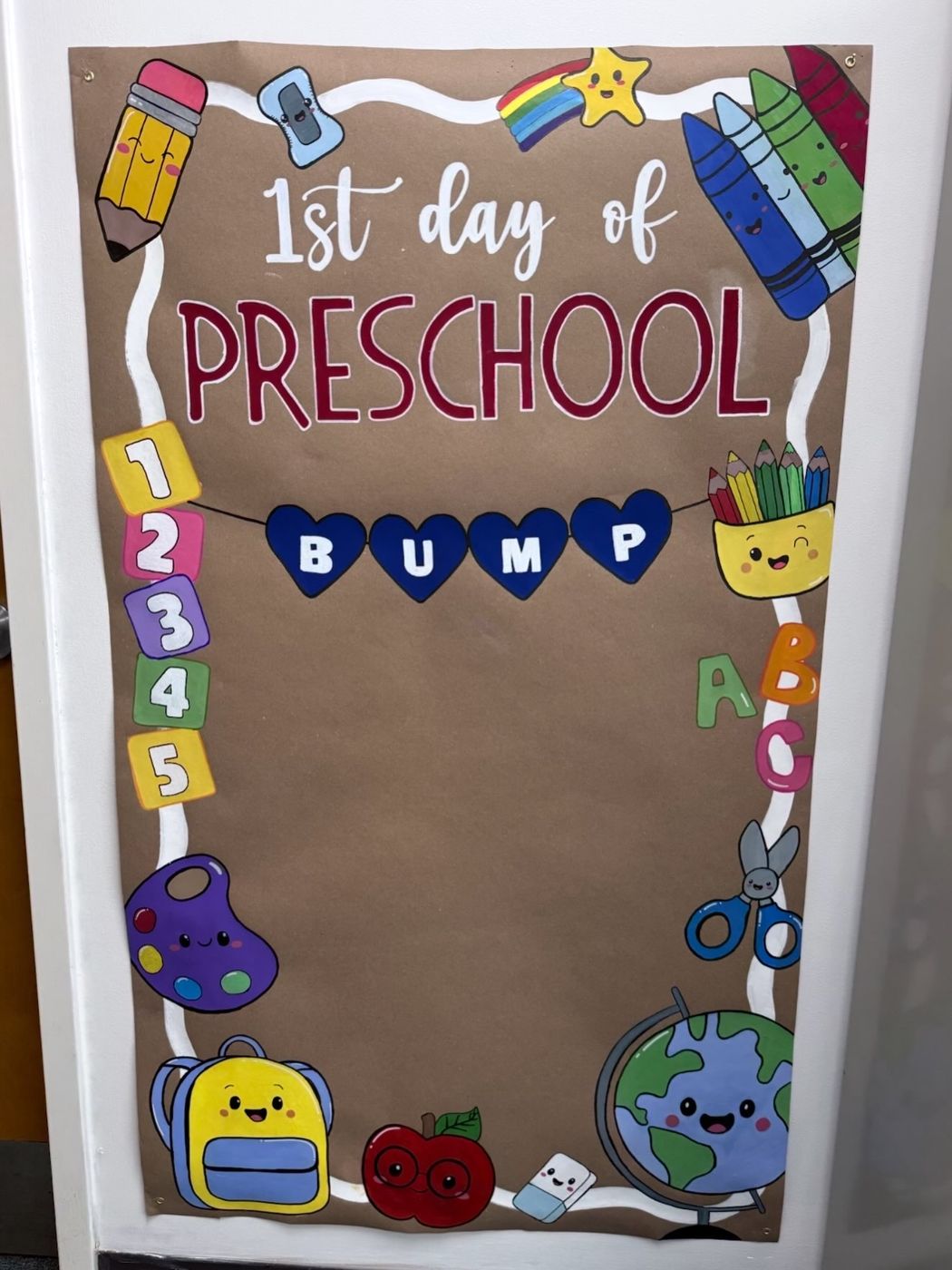First day of preschool photo board decorated with pencils, crayons, backpacks and a smiling globe